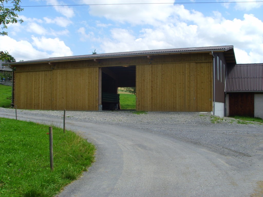 Eine große Holzscheune mit einer grünen Tür steht auf einem Kiesweg. Die Scheune hat ein Satteldach und ist von grünem Gras und ein paar Bäumen umgeben. Der Himmel darüber ist teilweise bewölkt.