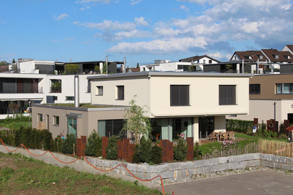 Modernes zweistöckiges Haus mit Flachdach, großen Fenstern und einer kleinen Terrasse, die von einem niedrigen Zaun umgeben ist. Das Haus liegt in einer Vorstadtgegend mit ähnlichen Gebäuden unter einem teilweise bewölkten Himmel. Grüner Rasen und Sträucher verschönern den Hof.