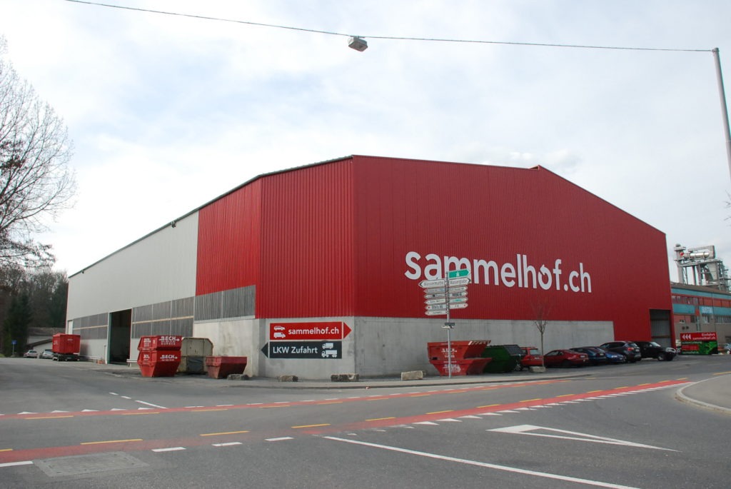 Grosses Industriegebäude mit rot-weisser Fassade und dem Schriftzug "sammelhof.ch". Daneben sind mehrere rote Container und parkende Autos zu sehen. Im Vordergrund sind Strassenschilder und eine Strassenecke zu sehen.