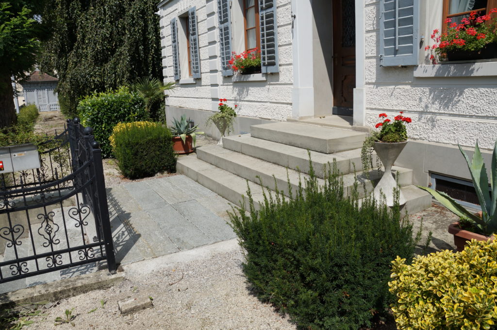 Ein weißer Hauseingang aus Stein mit grauen Fensterläden, roten Blumen in Töpfen auf den Stufen und viel Grün in der Umgebung. Auf der linken Seite ist teilweise ein schwarzes Eisentor sichtbar. Der Gehweg besteht aus Steinplatten.