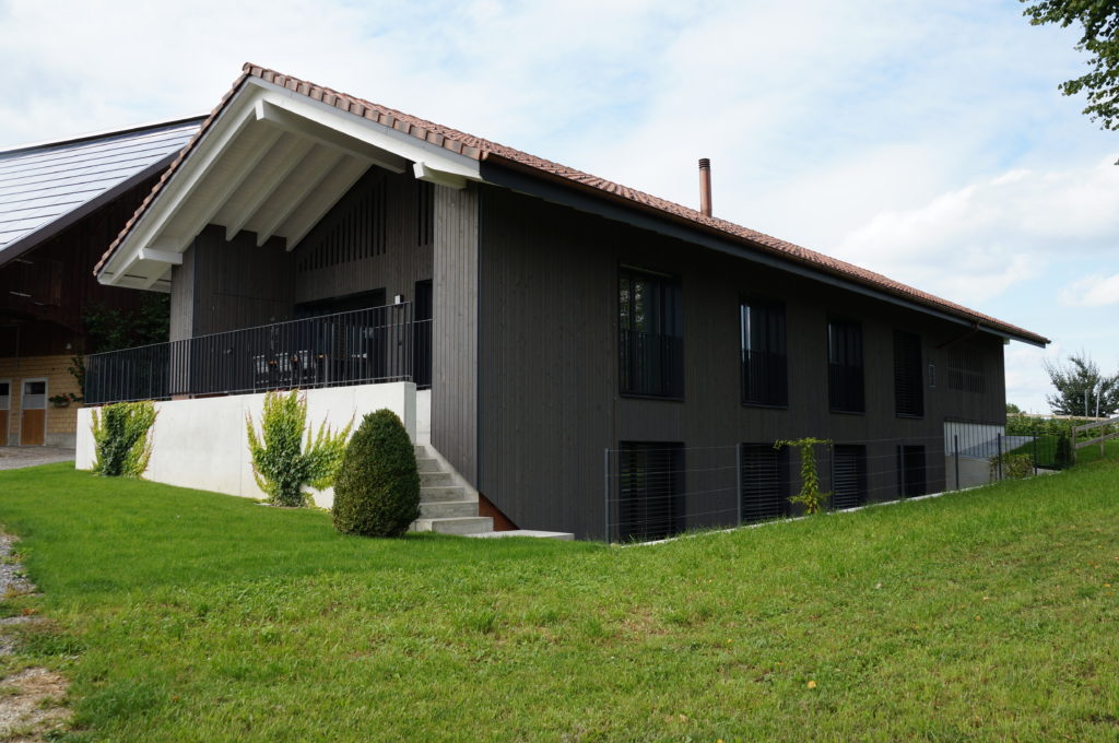 Einstöckiges Haus mit dunkler Holzfassade, rotem Ziegeldach und überdachter Veranda. Eine kleine Treppe führt zum Eingang. Das Haus ist von einem gepflegten Rasen und Sträuchern umgeben und der Himmel ist teilweise bewölkt.
