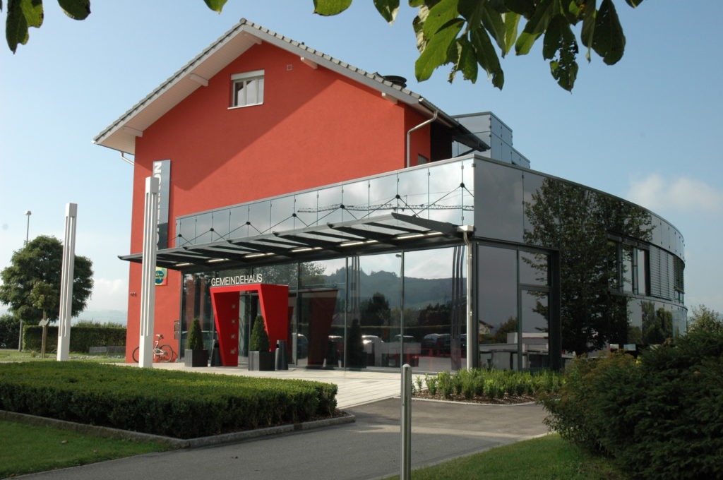 Ein modernes Gebäude mit roter Fassade und großen Glasfenstern, umgeben von Grün. Es hat ein schräges Dach und ein gläsernes Eingangsdach. Im Vordergrund und im Hintergrund sind Bäume und Büsche unter einem klaren blauen Himmel zu sehen.