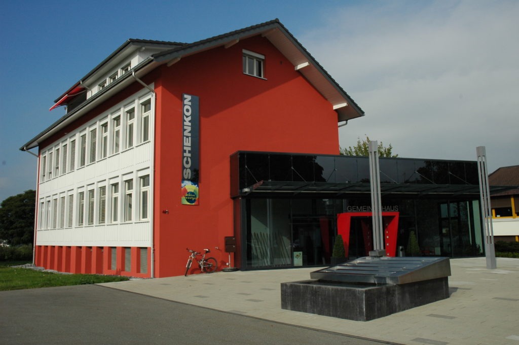 Ein modernes Gebäude mit rot-weißer Fassade und einem vertikalen Schild mit dem Wort „Schenkon“. Davor parkt ein Fahrrad, im Vordergrund steht eine schlanke Metallskulptur. Der Himmel ist bewölkt.