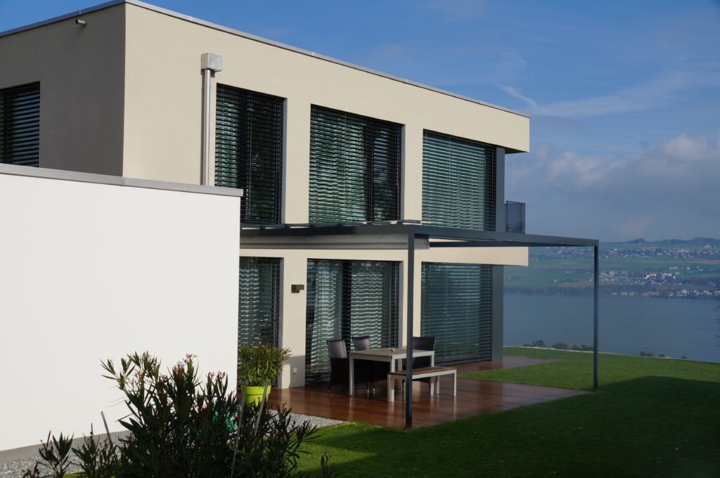 Modernes zweistöckiges Haus mit großen Fenstern und einer Außenterrasse mit Tisch und Stühlen. Mit Blick auf einen malerischen Blick auf einen See und ferne Hügel unter einem klaren blauen Himmel. Ein üppiger grüner Rasen umgibt das Haus.