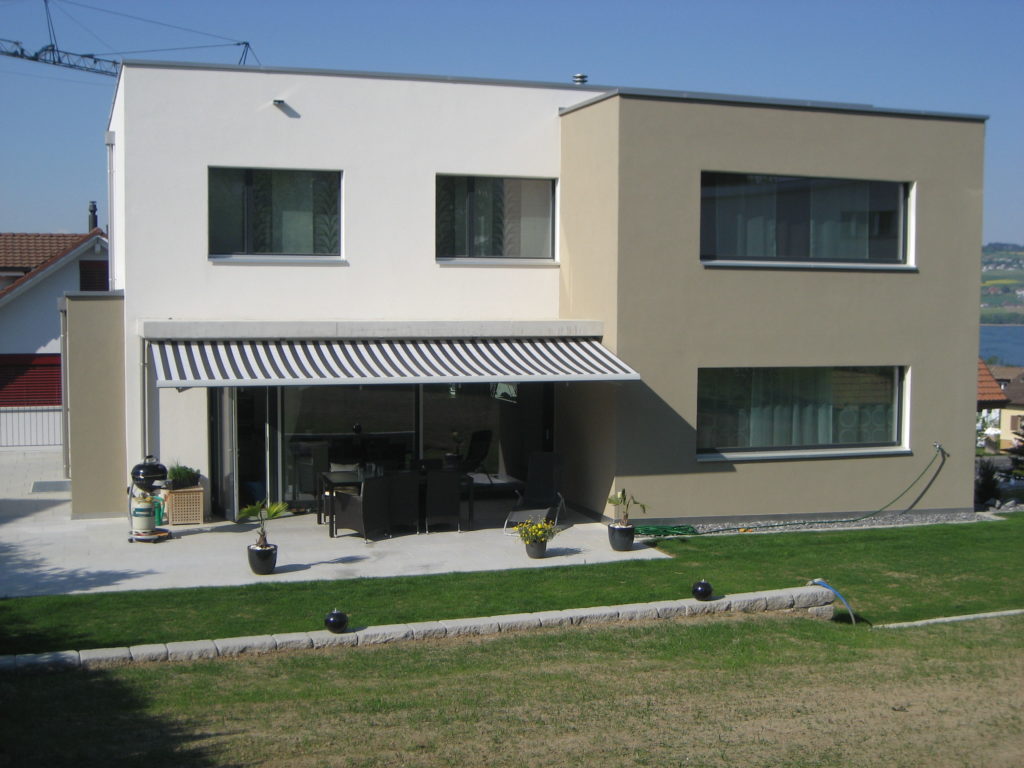 Ein modernes zweistöckiges Haus mit Flachdach und einer weiß-beigen Fassade. Der Hinterhof verfügt über eine Terrasse mit gestreifter Markise, Gartenmöbeln, Zierpflanzen und einem kleinen Rasen.