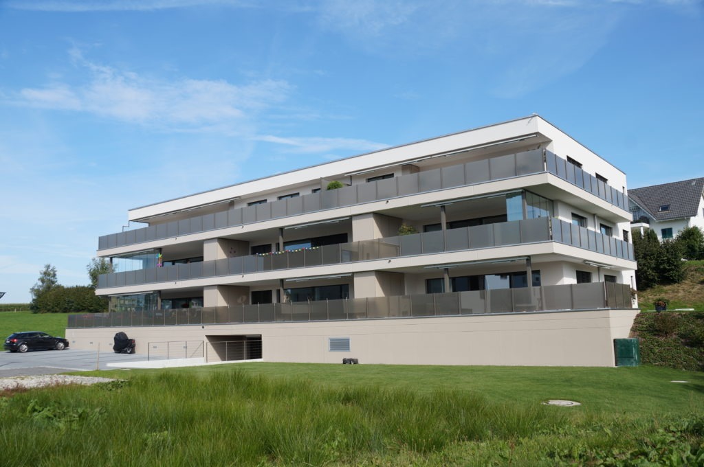 Ein modernes, dreistöckiges Wohngebäude mit Balkonen, umgeben von einem gepflegten Rasen. Der Himmel ist klar und blau und in der Einfahrt ist ein geparktes Auto zu sehen.
