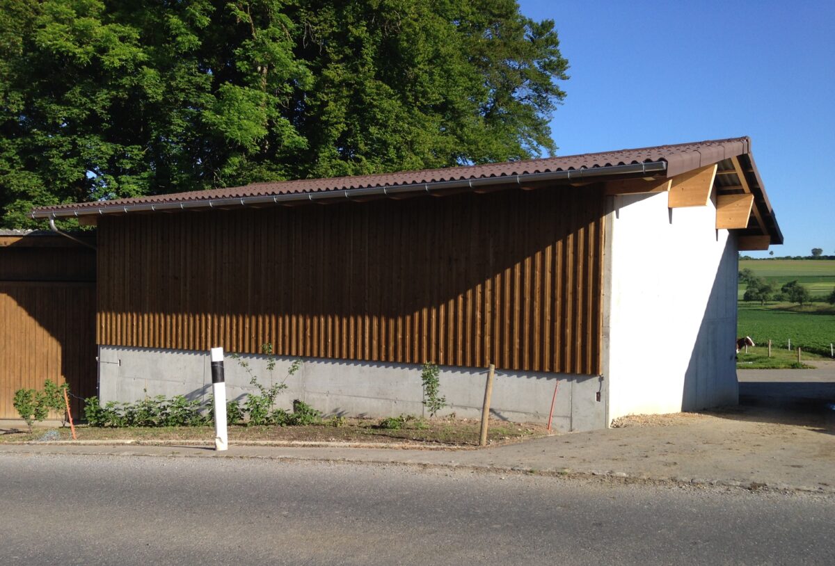 Ein Gebäude aus Holz und Beton mit schrägem Dach steht an einer Straße. Sonnenlicht wirft Schatten auf die Seite. In der Nähe der Straße steht ein Pfosten, im Hintergrund sind Bäume und grüne Felder unter einem klaren blauen Himmel zu sehen.