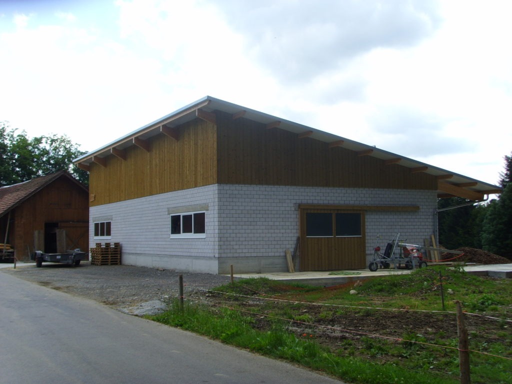 Eine moderne Scheune mit Schrägdach, Holzwänden im oberen Bereich und Betonwänden im unteren Bereich steht an einer Straße. Baumaterialien und Geräte liegen in der Nähe, im Vordergrund ist ein kleiner Fleck Gras und Erde zu sehen. Im Hintergrund sind Bäume zu sehen.