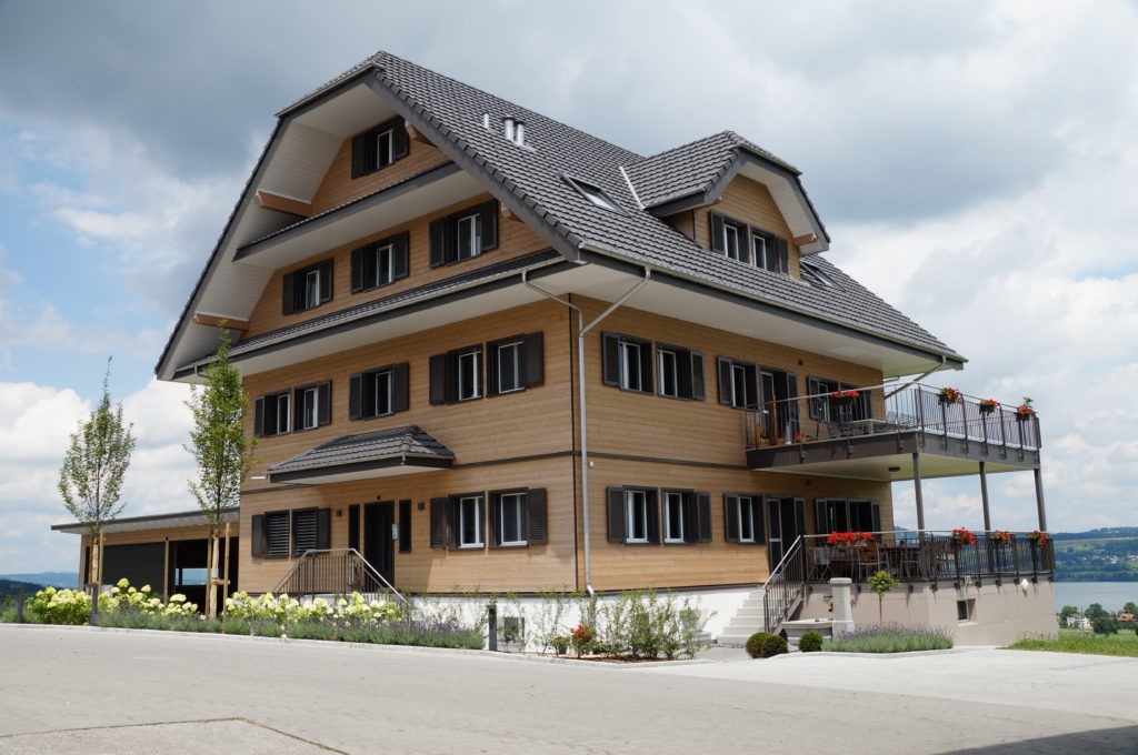 Großes, modernes Holzhaus mit steilem Satteldach, mehreren Fenstern und Balkonen, die mit roten Blumen geschmückt sind. Das Haus ist von einem gepflegten Garten umgeben und bietet Aussicht auf einen malerischen See im Hintergrund unter einem teilweise bewölkten Himmel.