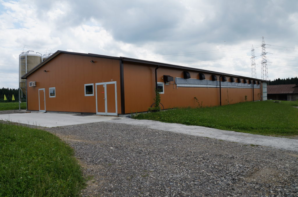 Ein großes braunes lagerhausähnliches Gebäude mit kleinen Fenstern und einer weißen Tür steht auf einem von Gras umgebenen Kiesweg. Im Hintergrund ist unter einem bewölkten Himmel ein Metallsilo zu sehen.