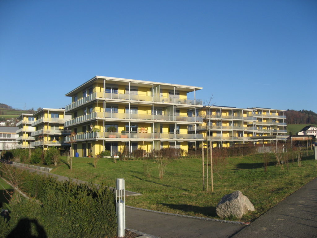 Vor einem klaren blauen Himmel erhebt sich ein modernes, mehrstöckiges Wohngebäude mit gelben Akzenten. Es ist von einem gepflegten Rasen und vereinzelten jungen Bäumen umgeben. Im Vordergrund befinden sich ein gepflasterter Gehweg und ein großer Felsen.