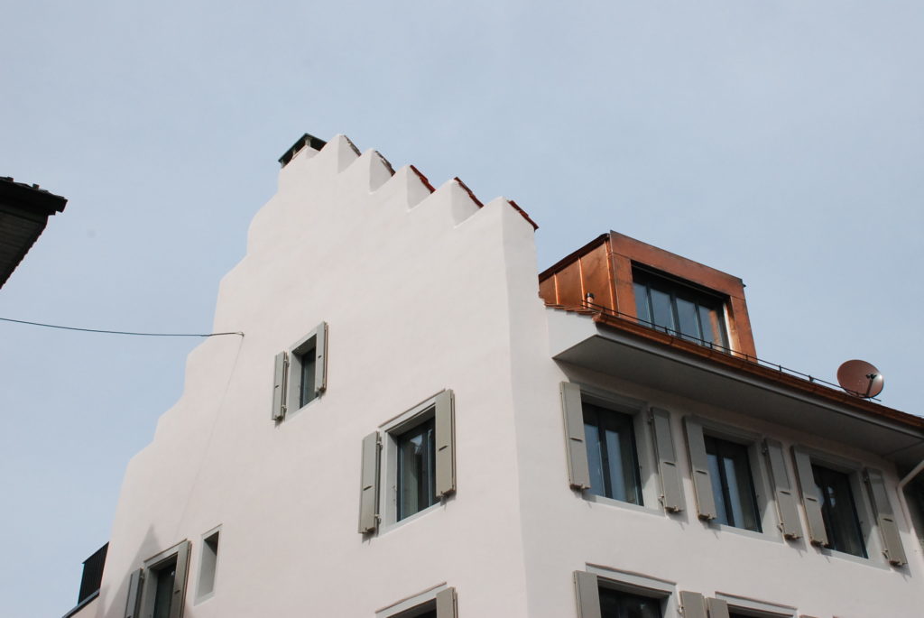 Ein modernes weißes Gebäude mit einzigartiger Stufenarchitektur auf einer Seite und mehreren Fenstern mit grauen Fensterläden. Das Dach hat einen rostfarbenen Abschnitt und eine Satellitenschüssel ist sichtbar. Der Himmel ist klar und blau.