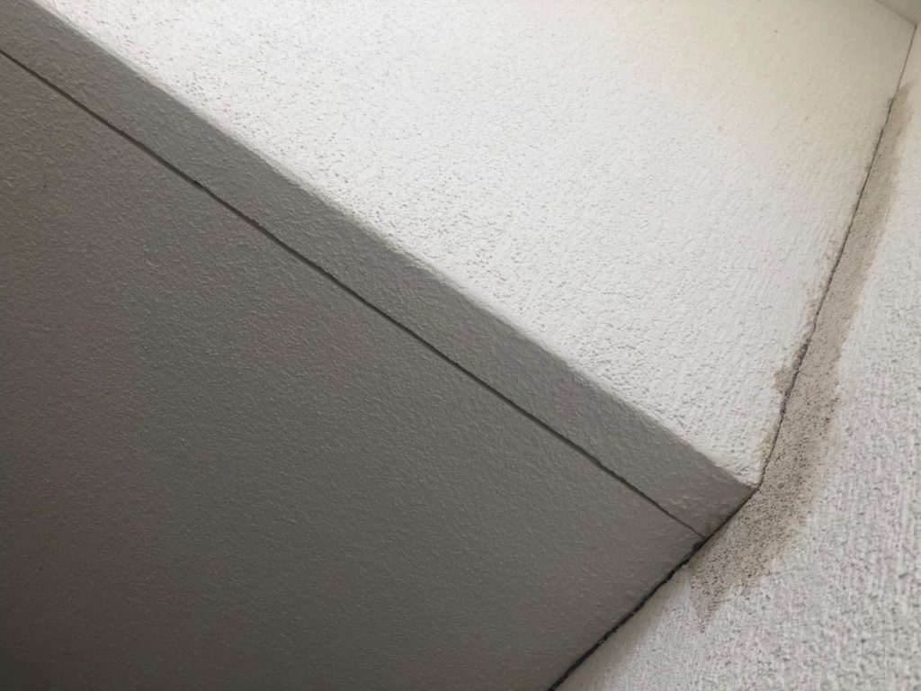 Nahaufnahme einer strukturierten weißen Decke mit einer sichtbaren Linie entlang der Kante, die eine Ecke zeigt, an der zwei Abschnitte aufeinandertreffen. Nahe der Kante auf der rechten Seite befindet sich ein kleiner Schmutz- oder Verfärbungsfleck.