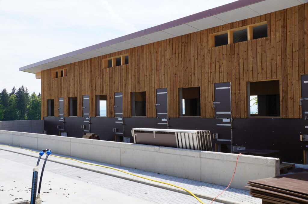 Ein im Bau befindliches modernes Gebäude mit einer Holzfassade und einer Reihe rechteckiger Fenster- und Türöffnungen. Am Sockel befindet sich eine Betonkonstruktion und überall sind Baumaterialien verstreut. Im Vordergrund liegt ein Schlauch auf dem Boden.