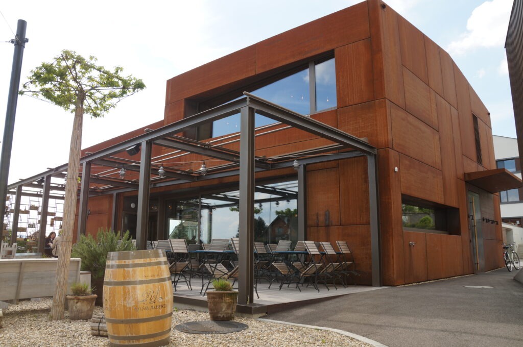 Modernes Café-Äußeres mit rostiger Metallfassade und Glasfenstern. Außensitzbereich unter einer Metallpergola mit mehreren Tischen und Stühlen. Ein Holzfass und kleine Pflanzen schmücken den Eingang. In der Nähe wächst ein schlanker Baum. Der Himmel ist teilweise bewölkt.