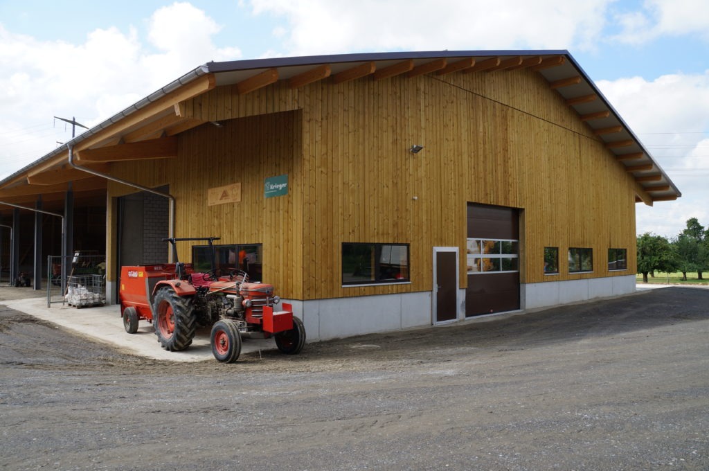 Eine große Holzscheune mit einem schrägen Metalldach steht auf einem Feldweg. Draußen steht neben einem offenen Garagentor ein roter Traktor. Der Himmel ist teilweise bewölkt und im Hintergrund sind Bäume zu sehen.