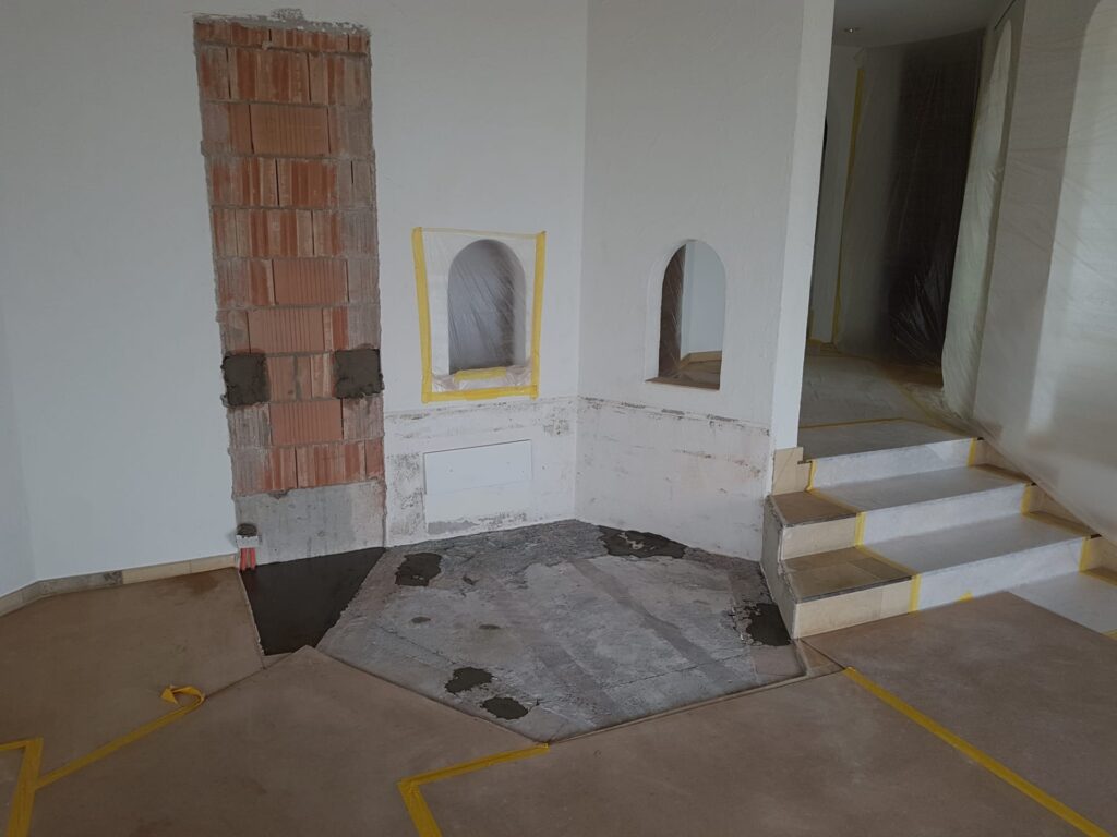 Eine Ecke eines Raums, der gerade renoviert wird. Sichtbare Ziegel und Putz. An einem Kaminbereich wird gearbeitet. Böden und Wände sind mit Schutzband versehen. Kleine gewölbte Nischen und eine Treppe sind sichtbar. In der Nähe befindet sich eine Plastikabdeckung.