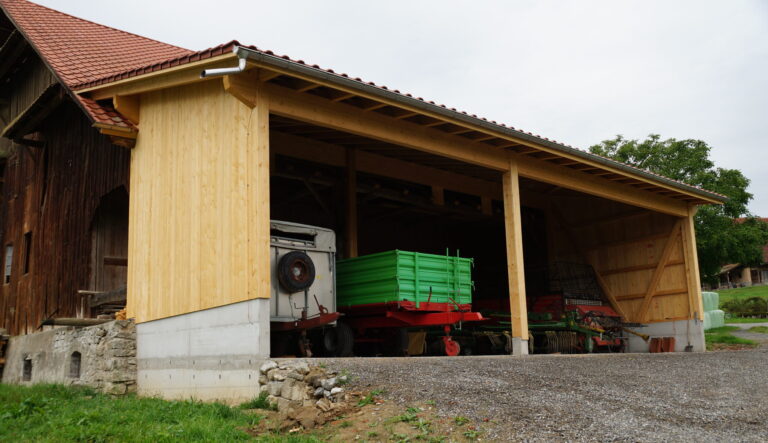 Ein Holzschuppen mit offener Vorderseite, in dem landwirtschaftliche Geräte untergebracht sind, darunter ein grüner Anhänger und verschiedene Maschinen. Das Gebäude hat ein rotes Ziegeldach und steht neben einem Kiesweg mit grünem Gras und Bäumen in der Nähe.