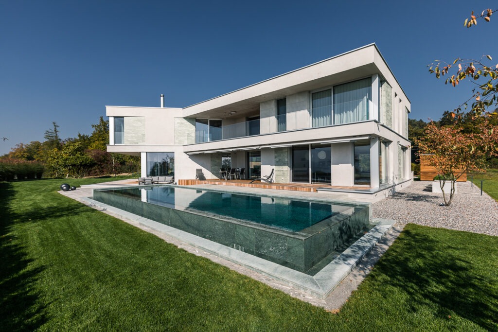 Modernes zweistöckiges Haus mit großen Fenstern, umgeben von Grün. Im Vordergrund befindet sich ein rechteckiger Swimmingpool, der von Gras umgeben ist. Der klare blaue Himmel im Hintergrund unterstreicht das schlichte, minimalistische Design des Gebäudes.