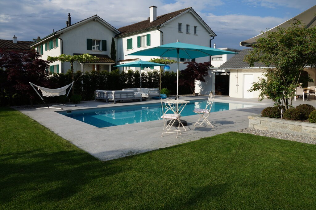 In einem Hinterhof steht ein rechteckiger Swimmingpool mit zwei weißen Stühlen und einem blauen Sonnenschirm daneben. In der Nähe ist eine Hängematte aufgehängt. Im Hintergrund sind unter einem bewölkten Himmel zwei Häuser mit grünen Fensterläden und gepflegten Büschen zu sehen.