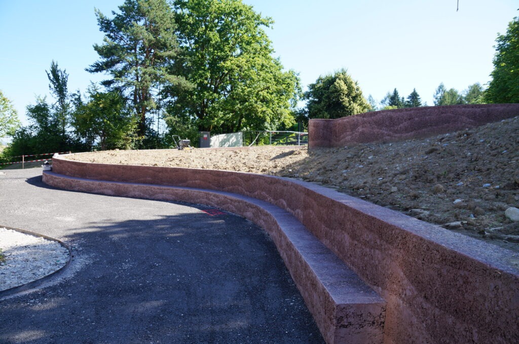 Gebogener Sitzbereich aus Beton mit Stützmauern in einer Parkanlage, umgeben von Bäumen und freiem Himmel. Der Bereich ist teilweise beschattet, mit einem abfallenden Grashügel über den Sitzplätzen.