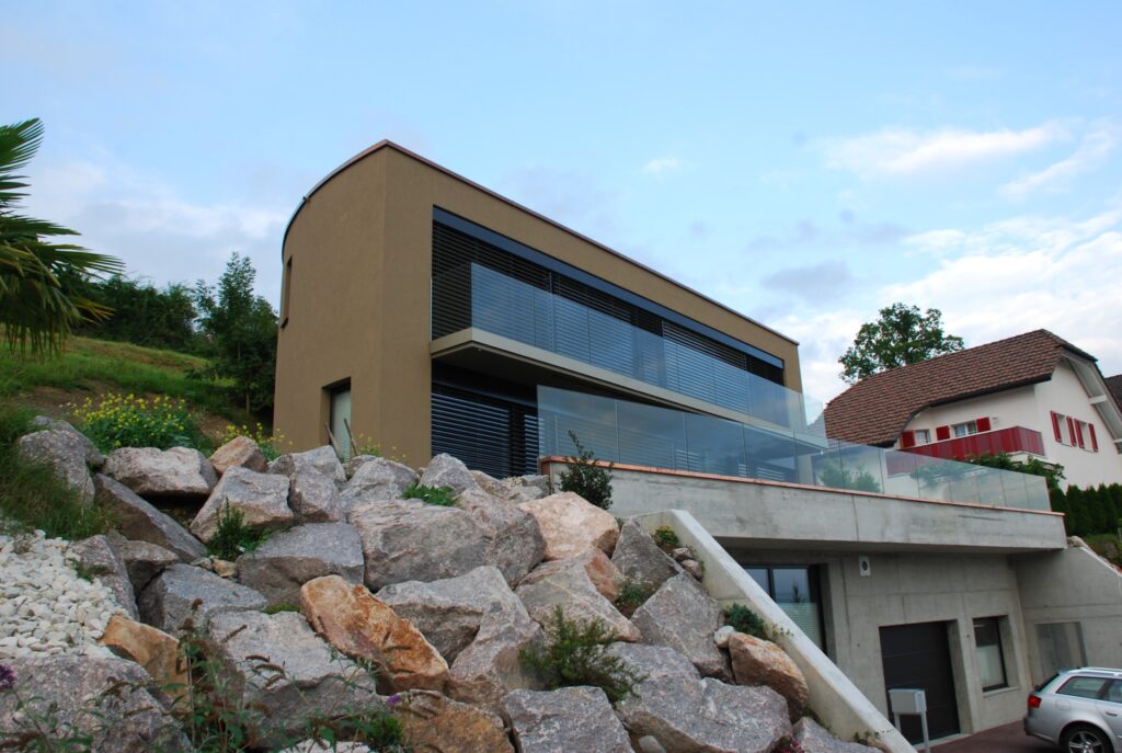 Modernes Haus mit großen Fenstern und Balkon, gelegen an einem Hügel mit Felsen und Grün. Das Gebäude hat ein schlichtes Design und grenzt an ein anderes traditionelles Haus. Ein Auto ist in der Nähe der Garage geparkt. Der Himmel ist teilweise bewölkt.