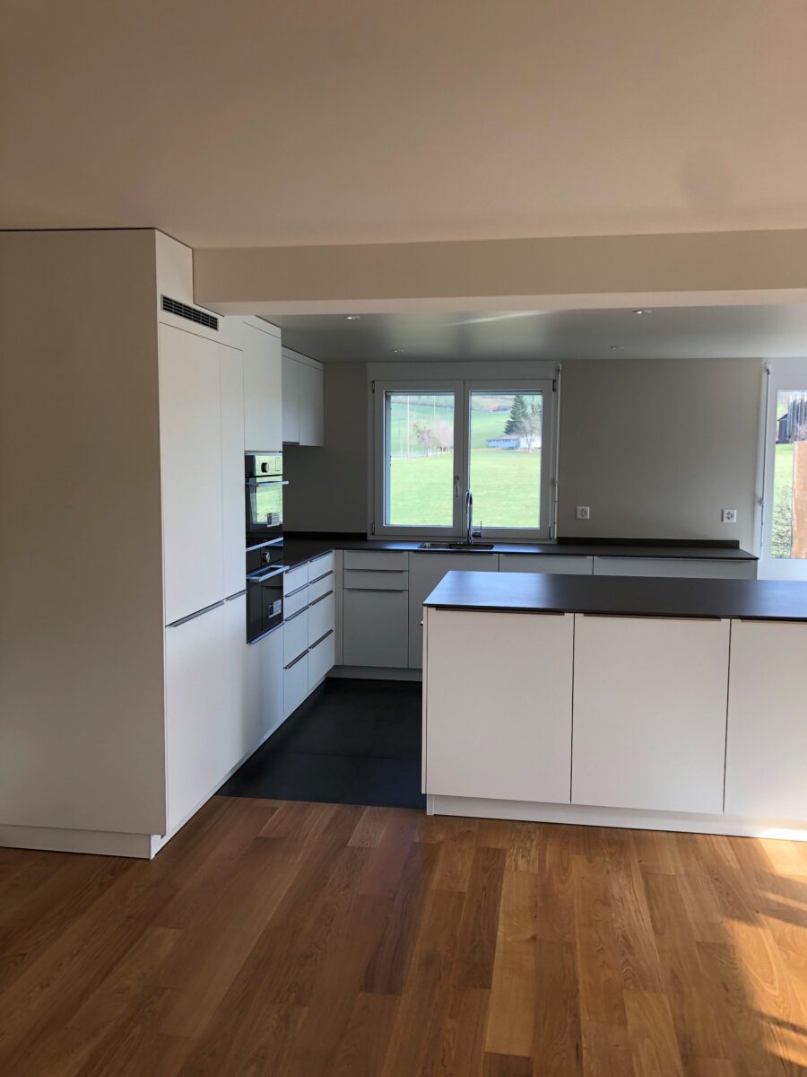 Moderne Küche mit weißen Schränken, einem eingebauten Ofen und einem Kochfeld. Der Raum hat Holzfußboden und ein Fenster, das für Tageslicht sorgt und auf einen grünen Rasen blickt. Die Arbeitsplatten sind dunkel und bieten einen eleganten Kontrast zu den weißen Schränken.