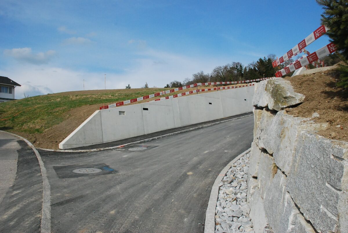 Eine neu asphaltierte kurvenreiche Straße mit weißen und roten Bauzäunen auf der rechten Seite, angrenzend an einen grasbewachsenen Abhang. Eine Steinmauer folgt der Kurve der Straße und in der Ferne ist unter einem teilweise bewölkten blauen Himmel ein Gebäude zu erkennen.