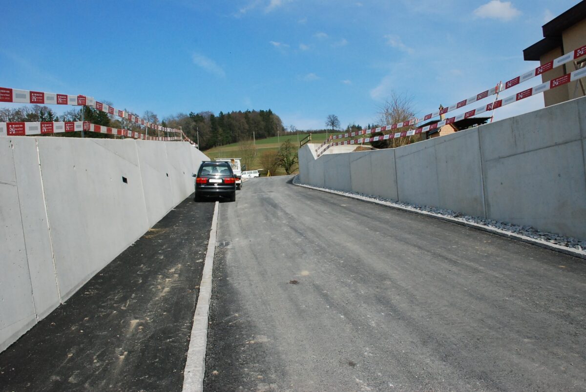 Ein Auto steht auf einer asphaltierten Straße, die von Betonmauern flankiert wird, die sich im Bau befinden. An den Oberkanten der Mauern sind rote und weiße Leitplanken angebracht. Im Hintergrund sind Bäume und ein klarer blauer Himmel zu sehen.