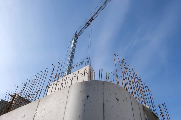 Baustelle mit teilweise fertiggestellter Betonkonstruktion und freiliegendem Bewehrungsstahl. Ein hoher Kran ragt in den blauen Himmel und deutet auf laufende Bauarbeiten hin. Die Szene lässt auf eine Entwicklung oder einen laufenden Bau schließen.