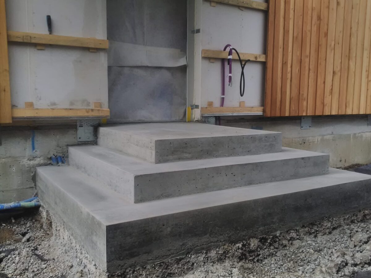 Drei neu errichtete Betonstufen führen zu einer Tür. Die Stufen sind von einer Kiesfläche umgeben und das Gebäude verfügt über unfertige Wände mit freiliegendem Holz und Isoliermaterialien.
