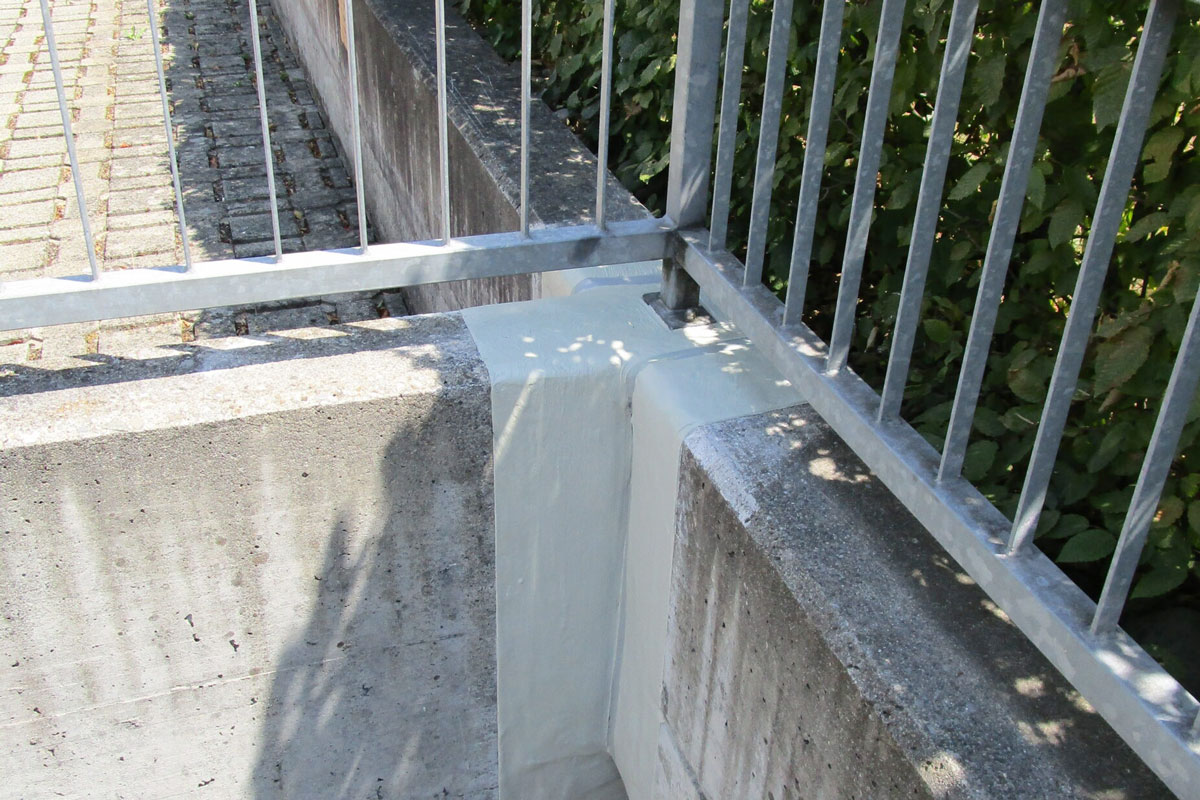 Ein Metallgeländer an der Ecke einer niedrigen Betonmauer, an deren Fuge ein hellerer Betonfleck zu sehen ist. Auf die Oberfläche werden Schatten von Blättern geworfen und links ist teilweise ein Kopfsteinpflasterweg zu sehen.