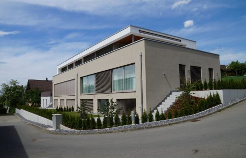 Modernes zweistöckiges Haus mit großen Fenstern und horizontalen Holzlamellen, umgeben von einer gepflegten Hecke. Die Außenfassade ist beige mit einem Flachdach. Eine weiße Treppe führt zum Eingang. Der Himmel ist klar mit einigen Wolken.
