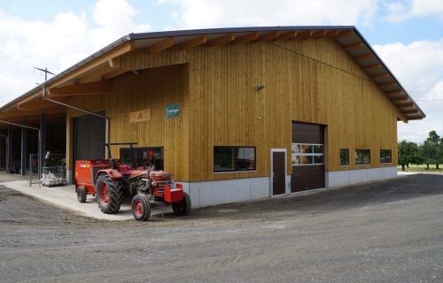 Eine große Holzscheune mit einem schrägen Metalldach steht auf einem Feldweg. Draußen steht neben einem offenen Garagentor ein roter Traktor. Der Himmel ist teilweise bewölkt und im Hintergrund sind Bäume zu sehen.