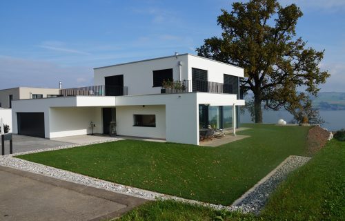 Modernes zweistöckiges weißes Haus mit schwarzen Fensterläden. Es verfügt über ein Flachdach und eine große Terrasse. Das Haus ist von Grün umgeben, darunter ein Baum auf der rechten Seite. Eine Auffahrt führt zu einer Garage auf der linken Seite. Im Hintergrund ist ein Blick auf einen See oder das Meer zu sehen.