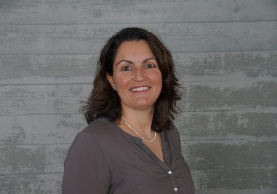 Eine Person mit schulterlangem braunem Haar lächelt und trägt ein graues Hemd und eine Perlenkette. Sie steht vor einer strukturierten grauen Wand.