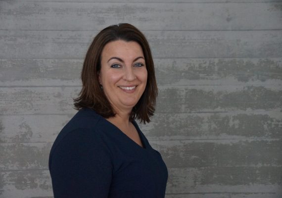 Frau mit schulterlangem braunem Haar und marineblauem Oberteil lächelt, während sie vor einer strukturierten grauen Wand steht.