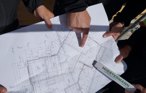Drei Personen untersuchen einen detaillierten Bauplan. Eine Person hält das Papier mit einem Lineal, während eine andere auf bestimmte Abschnitte des Plans zeigt. Der Bauplan enthält verschiedene Maße und Anmerkungen.