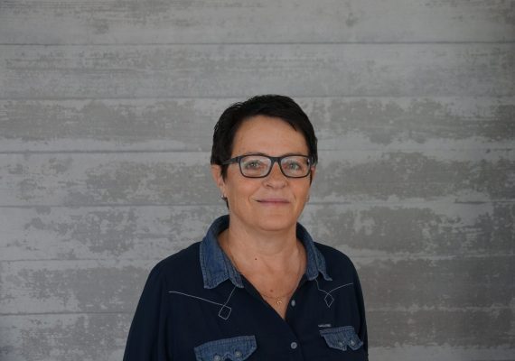 Eine Person mit kurzen dunklen Haaren und Brille trägt ein dunkelblaues Hemd mit Jeansakzenten. Sie lächelt und steht vor einem grauen, strukturierten Hintergrund.